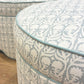 Two Highland House Suzie Scalloped Ottomans, upholstered in pale blue and white patterned fabric with blue piping, offer a stylish footrest or versatile accent on a wooden floor in a bright room.