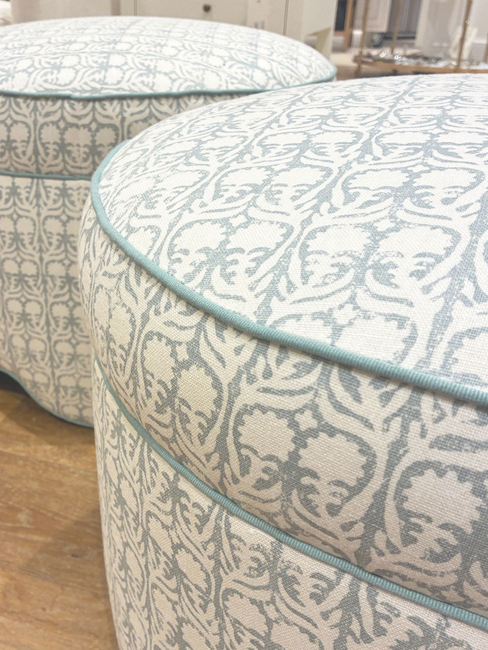 Two Highland House Suzie Scalloped Ottomans, upholstered in pale blue and white patterned fabric with blue piping, offer a stylish footrest or versatile accent on a wooden floor in a bright room.