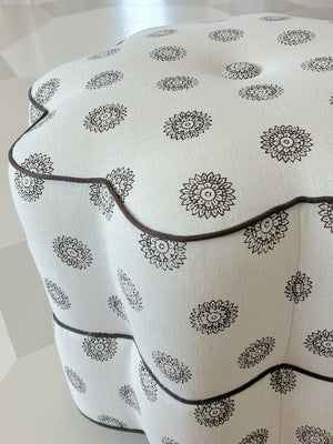 A Bunny Williams Stella Stool ottoman with a white scalloped edge, dark floral patterns, and dark piping sits on a light checkered floor.