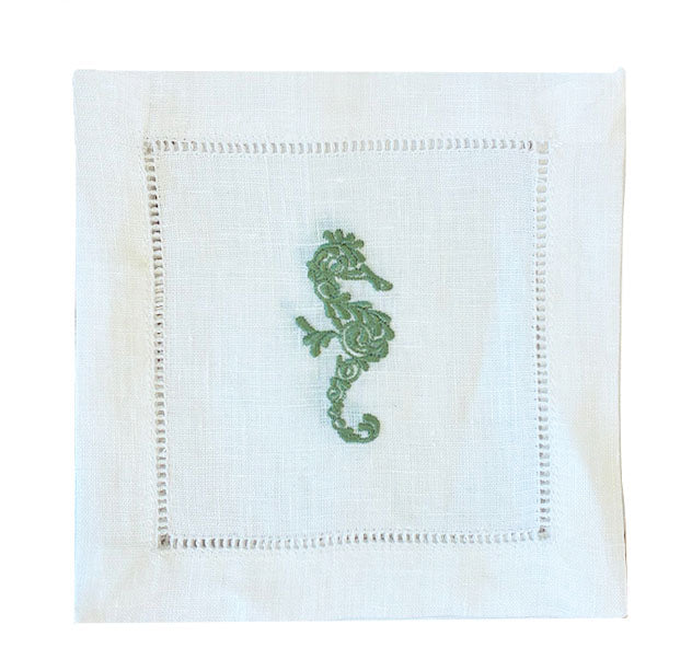 The Coastal Cocktail Napkin Set by Pink House Mustique features white linen napkins with a decorative cutwork border and an embroidered green seahorse, perfect for bringing coastal charm to your table.