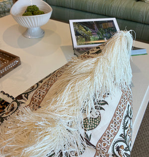 Close-up of the Amazonas Mystique Pillow by Associated Design, featuring beige fringe and patterned fabric on a coffee table, with a bowl, book, and tray in the background.
