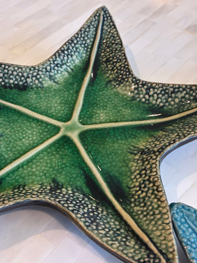 A close-up of a textured, deep green starfish-shaped plate from the Green Starfish Dish Collection by Van Cleve Collection, displayed on a light-colored tiled surface.