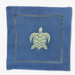 The Coastal Cocktail Napkin Set by Pink House Mustique features coastal blue linen napkins with a light green embroidered turtle in the center and decorative edge stitching.