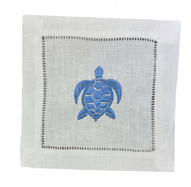 The Coastal Cocktail Napkin Set by Pink House Mustique features white linen napkins with a decorative border and a blue embroidered turtle, ideal for bringing a coastal vibe to your table.