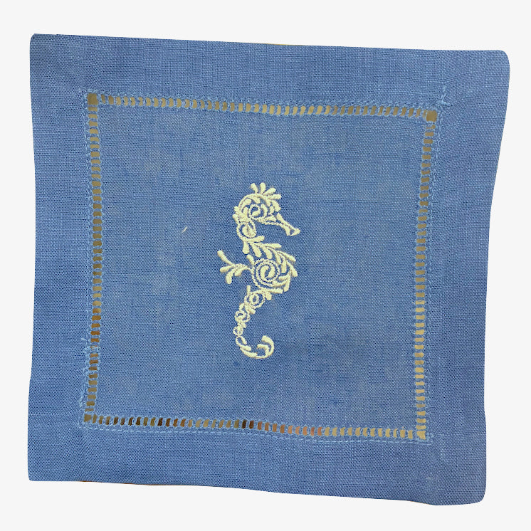 The Coastal Cocktail Napkin Set by Pink House Mustique features blue linen napkins with a decorative yellow embroidered seahorse at the center and elegant openwork stitching details.