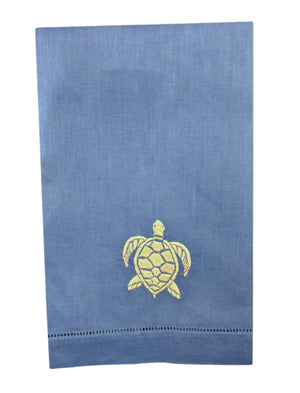 The Coastal Linen Hand Towel Collection by Pink House Mustique features a blue hand towel with a yellow embroidered sea turtle near the bottom center, adding seaside charm and coastal style to your table setting.