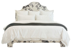 The EJ Schrader Pyne Hollyhock King Bed, featuring botanical patterns on the headboard and base, is styled with white bedding, multiple white pillows, and a dark textured throw blanket folded at the foot.
