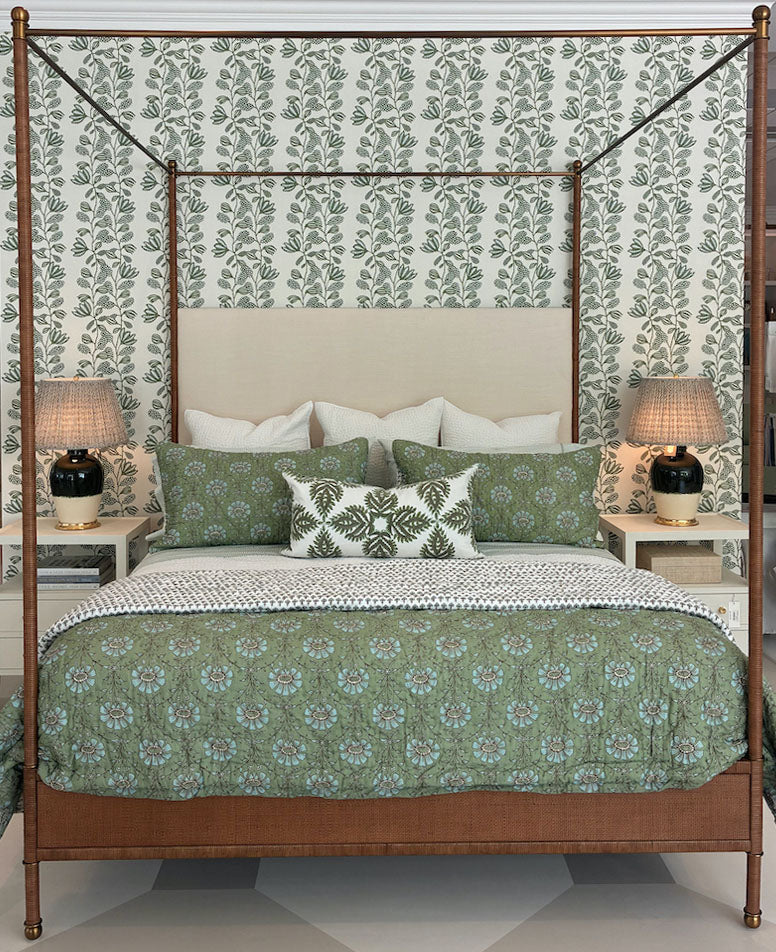 A neatly made Julian Chichester Rita Cane Wrap Four Poster Bed with green patterned bedding and pillows, flanked by two lamps on white nightstands against a leafy wallpaper background.