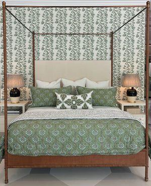 A neatly made Julian Chichester Rita Cane Wrap Four Poster Bed with green patterned bedding and pillows, flanked by two lamps on white nightstands against a leafy wallpaper background.