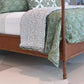 A close-up of the Julian Chichester Rita Cane Wrap Four Poster Bed, featuring a wooden frame with cane detailing, dressed in white and green floral bedding and patterned sheets, displayed on a geometric-patterned floor.