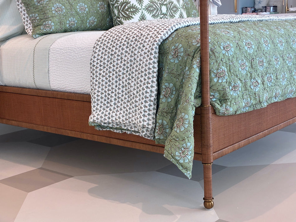 A close-up of the Julian Chichester Rita Cane Wrap Four Poster Bed, featuring a wooden frame with cane detailing, dressed in white and green floral bedding and patterned sheets, displayed on a geometric-patterned floor.
