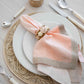 A neatly set table includes a woven placemat, white plates, cream cutlery, and a peach and beige napkin adorned with the Kim Seybert Playa Napkin Ring and organic bead baubles.