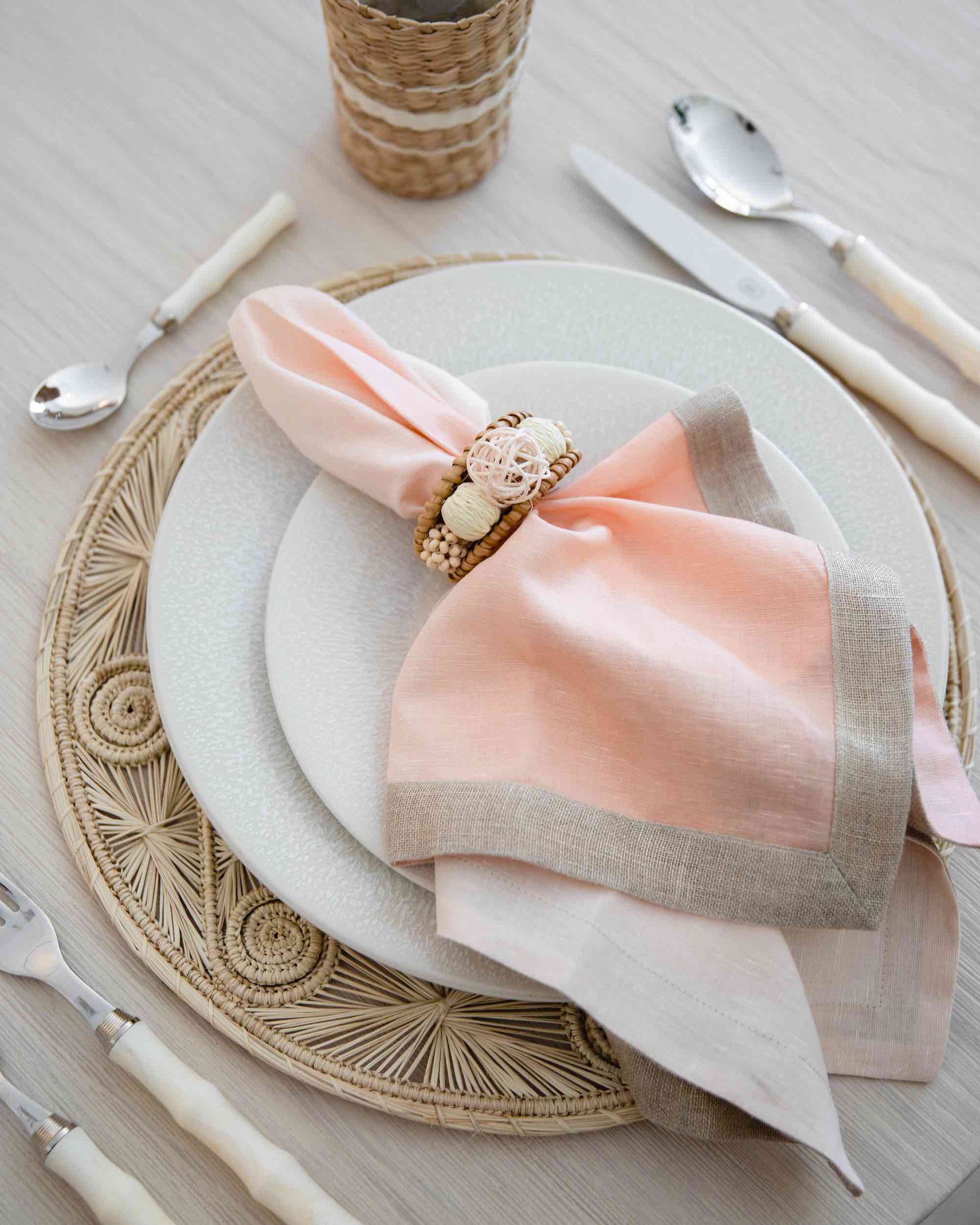 A neatly set table includes a woven placemat, white plates, cream cutlery, and a peach and beige napkin adorned with the Kim Seybert Playa Napkin Ring and organic bead baubles.