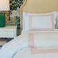 A neatly made bed with white Matouk HIVE Exclusive Rovella Bedding Collection in Salmon, featuring Egyptian cotton percale and pink geometric borders, sits by a beige headboard, white nightstand, green lamp, and floral wallpaper.