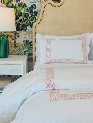 A neatly made bed with white Matouk HIVE Exclusive Rovella Bedding Collection in Salmon, featuring Egyptian cotton percale and pink geometric borders, sits by a beige headboard, white nightstand, green lamp, and floral wallpaper.