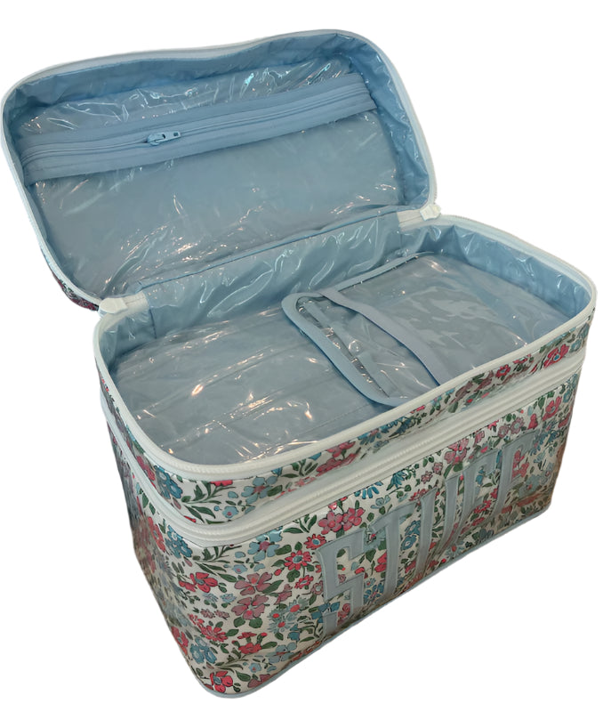 The Walker Valentine Walton Travel Case, featuring a floral pattern and double zippers, is open on a woven surface to reveal spacious, clear plastic-lined compartments.