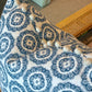 A close-up of the Tassel Inverted Medallion Navy Pillow by Associated Design, featuring handmade blue floral designs and decorative buttons, resting on a textured beige carpet near wooden and cane furniture.