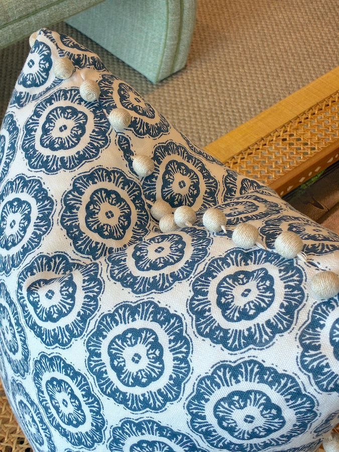 A close-up of the Tassel Inverted Medallion Navy Pillow by Associated Design, featuring handmade blue floral designs and decorative buttons, resting on a textured beige carpet near wooden and cane furniture.