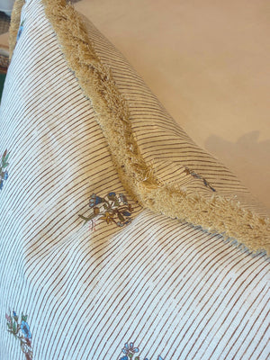 A close-up of the Flax Field Pinstripe Brush Fringe Pillow by Hive Custom Pillows, featuring gold fringe trim and a striped floral pattern, crafted from premium materials and set against a plain beige background.