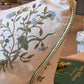The Flax Field Flower with Leaf Trim Pillow by Hive Custom Pillows features a floral pattern and decorative trim, shown on a sofa near green books in a bright room, showcasing its luxurious materials.
