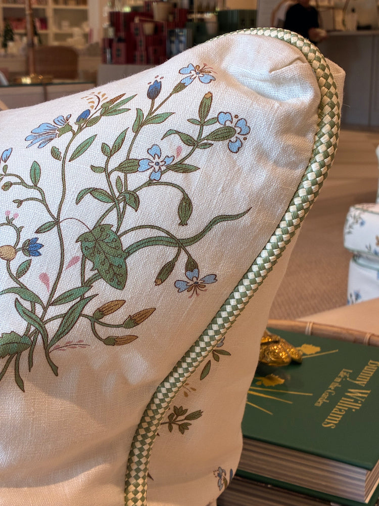 The Flax Field Flower with Leaf Trim Pillow by Hive Custom Pillows features a floral pattern and decorative trim, shown on a sofa near green books in a bright room, showcasing its luxurious materials.