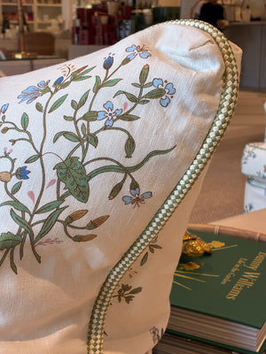 The Flax Field Flower with Leaf Trim Pillow by Hive Custom Pillows features a floral pattern and decorative trim, shown on a sofa near green books in a bright room, showcasing its luxurious materials.