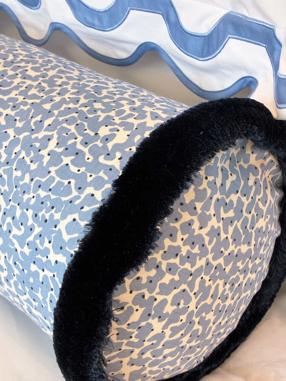 The Korallo Periwinkle Bolster Pillow by Hive Custom Pillows features a cylindrical blue and white pattern with black trim, shown in front of a white pillow with a blue wavy border.