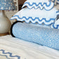 A bed with white linens, blue scalloped edges, a Korallo Periwinkle Bolster Pillow by Hive Custom Pillows in blue and white print, and a lamp with a matching patterned shade—all crafted from the finest materials.