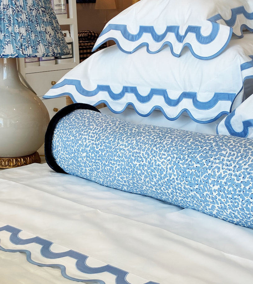 A bed with white linens, blue scalloped edges, a Korallo Periwinkle Bolster Pillow by Hive Custom Pillows in blue and white print, and a lamp with a matching patterned shade—all crafted from the finest materials.