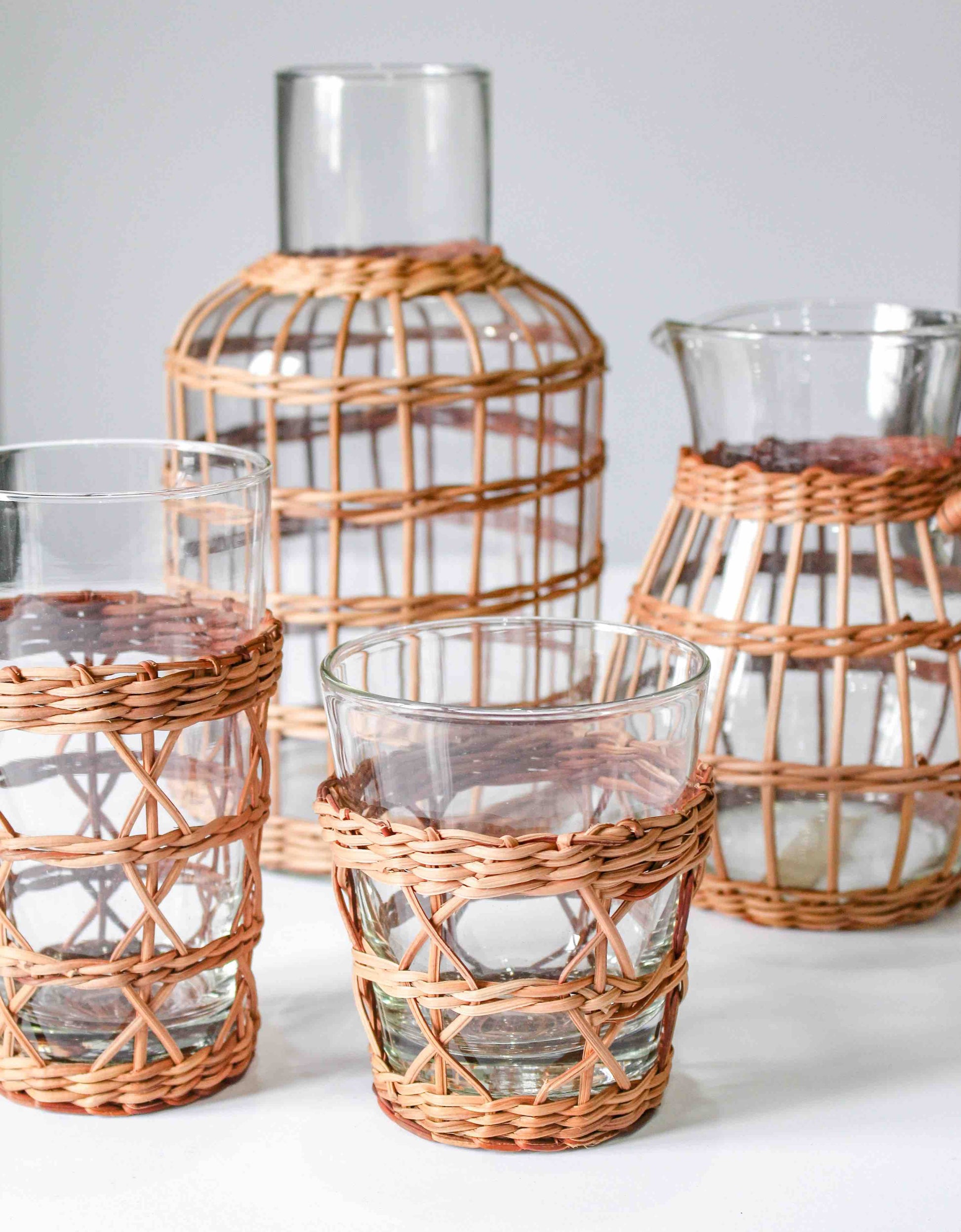 The Be Home Seagrass Rattan Cage Tumbler Collection features four glass tumblers wrapped in decorative woven wicker, combining rustic charm and modern convenience, shown on a plain white background.