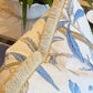 Close-up of Hive Custom Pillows’ Enchanted Garden Pillow, featuring a blue botanical print and beige fringe, made from premium materials, shown on furniture with a plant in the background.