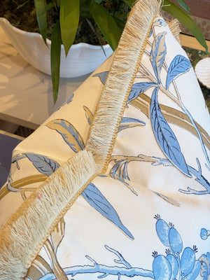 Close-up of Hive Custom Pillows’ Enchanted Garden Pillow, featuring a blue botanical print and beige fringe, made from premium materials, shown on furniture with a plant in the background.