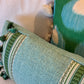 Two Pyla Lagon Pillows by Hive Custom Pillows, featuring green and white patterns with tassels, rest on a light-colored textured fabric surface.