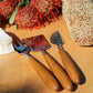 Three cheese knives from the Be Home Stainless Steel and Teak Cheese Set rest on a wooden surface near a loaf of seeded bread, a wedge of cheese, and a vibrant red flowering plant.