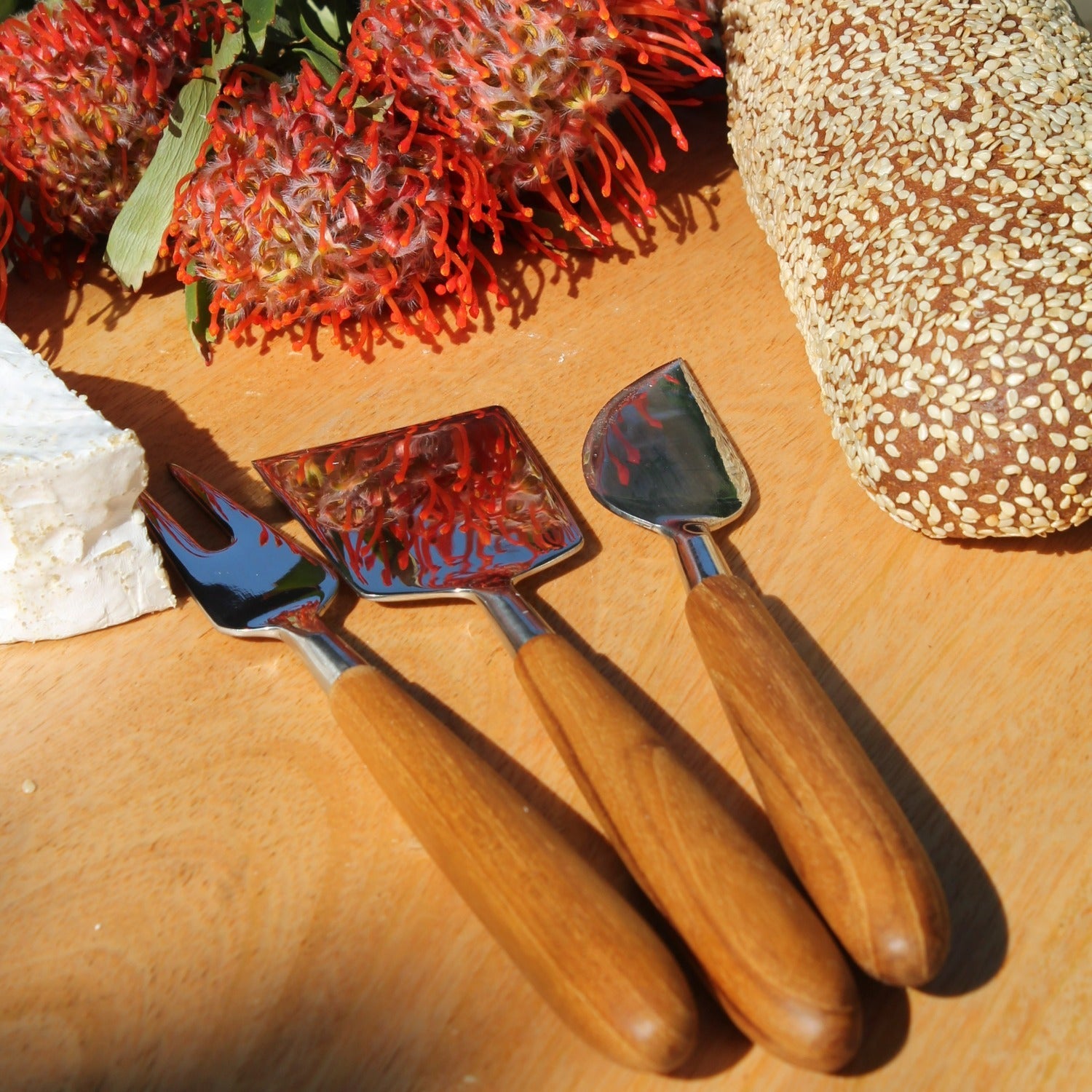 Three cheese knives from the Be Home Stainless Steel and Teak Cheese Set rest on a wooden surface near a loaf of seeded bread, a wedge of cheese, and a vibrant red flowering plant.