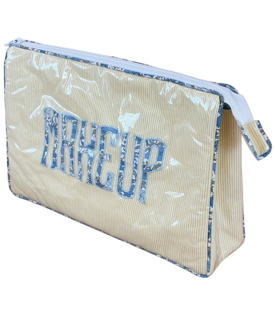 The Walker Valentine Large Helen Cosmetic Case, featuring "MAKEUP" in bold blue on a light yellow and white striped design, is a roomy rectangular zippered bag—ideal for organizing cosmetics at home or while traveling.