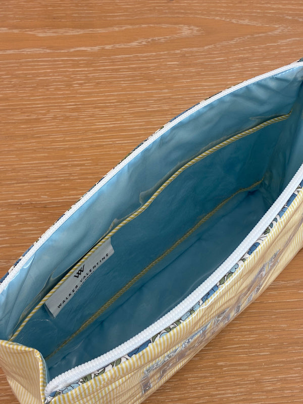 The Large Helen Cosmetic Case by Walker Valentine features a blue interior, multiple inner pockets, a "MAKEUP" label inside, and sits on a wooden surface—ideal for organizing cosmetics.