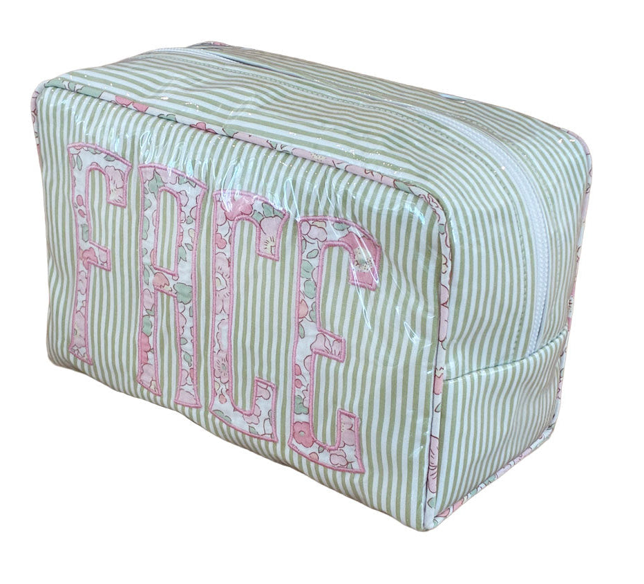 The Walker Valentine Large Milan Cosmetic Case, "FACE," features green and white stripes, a zip closure, and bold pink floral letters—ideal as a travel makeup bag or cosmetic organizer.