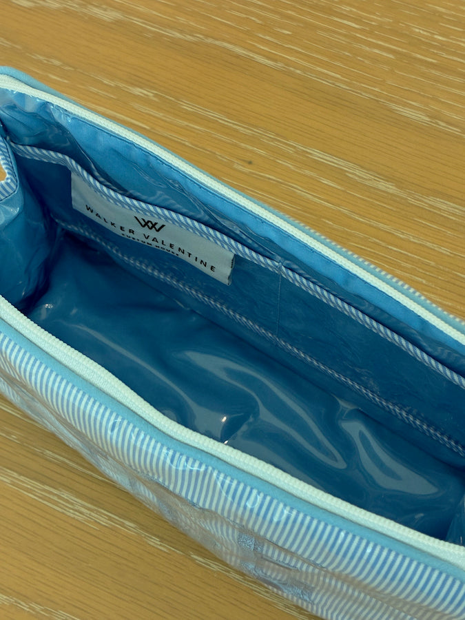 The Small Helen Cosmetic Case "FACE" by Walker Valentine features blue and white stripes, a zippered closure, blue interior, and label—perfect for travel and cosmetics. Shown here on a wooden surface.