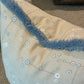 Close-up of the Klimt Grid Pillow with Sky Brush Fringe by Associated Design, featuring a beige base, blue fringe, and circular pattern, resting on a textured brown carpet with another cushion partially visible behind.