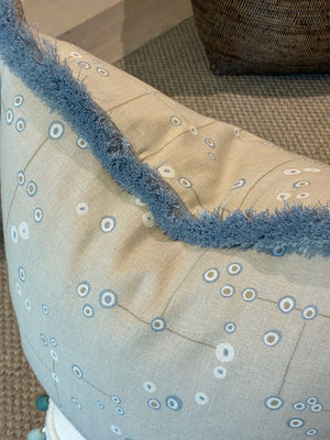 Close-up of the Klimt Grid Pillow with Sky Brush Fringe by Associated Design, featuring a beige base, blue fringe, and circular pattern, resting on a textured brown carpet with another cushion partially visible behind.