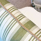 The Tasca Stripe Pillow with Burlap Cord Trim by Hive Custom Pillows features green and white stripes and tan cord trim, displayed on a white surface with a textured beige carpet and tasseled handmade pillow in the background.