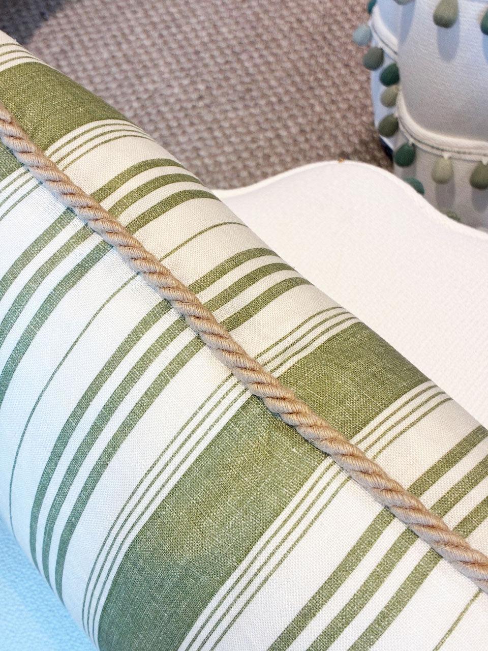 The Tasca Stripe Pillow with Burlap Cord Trim by Hive Custom Pillows features green and white stripes and tan cord trim, displayed on a white surface with a textured beige carpet and tasseled handmade pillow in the background.