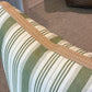 A close-up of the Hive Custom Pillows Tasca Stripe Box Edge Pillow with Burlap Border, featuring green and white stripes and tan trim, displayed on a beige carpet near a wicker basket and wooden table.