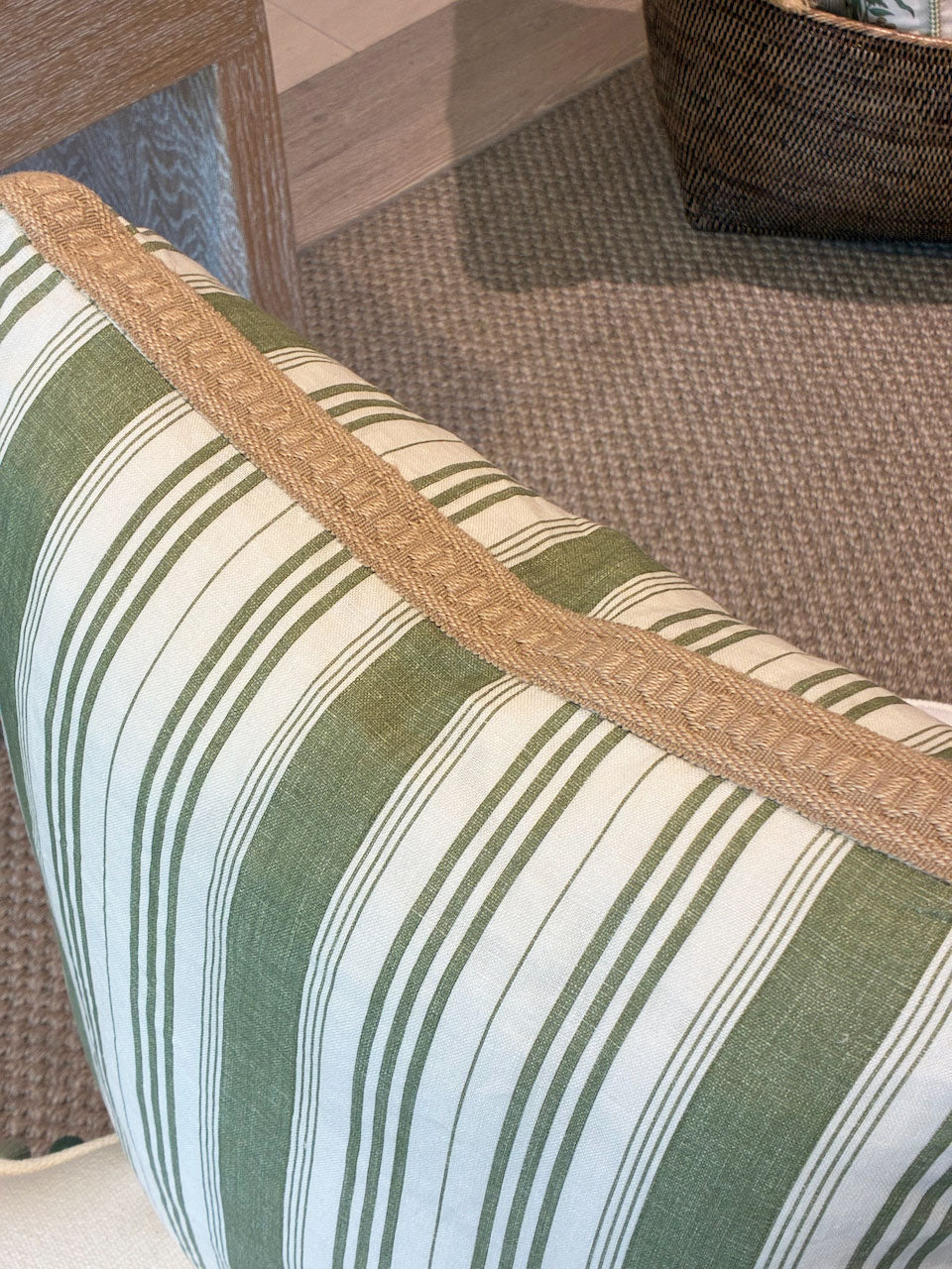 A close-up of the Hive Custom Pillows Tasca Stripe Box Edge Pillow with Burlap Border, featuring green and white stripes and tan trim, displayed on a beige carpet near a wicker basket and wooden table.
