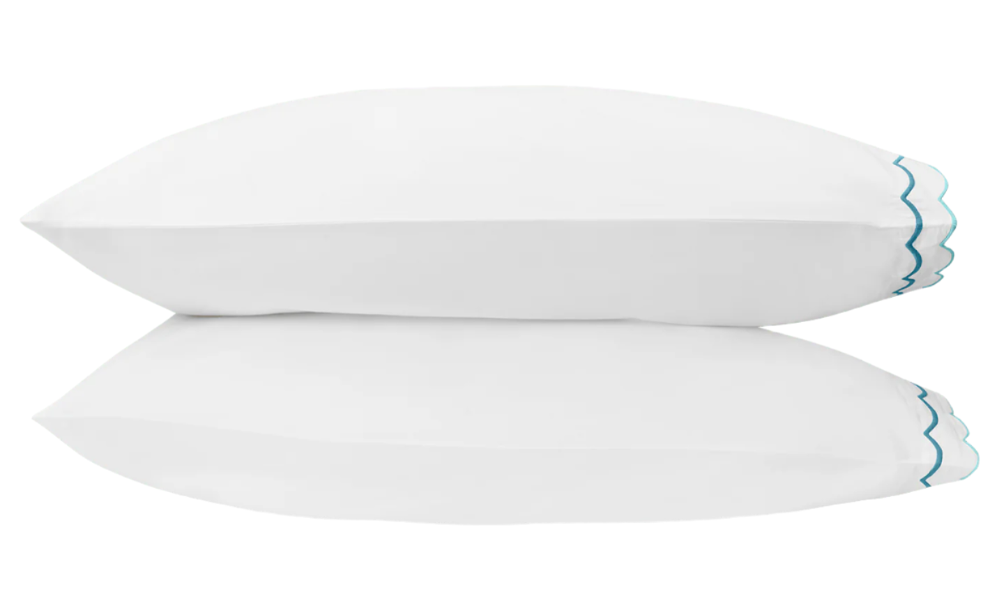 The Matouk India Bedding Collection, Cerulean—a white pillow with blue wave-like embroidery—reflects on a glossy surface.