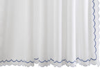 The India Pique Shower Curtain Collection by Matouk features a white cotton blend curtain with a subtle diamond pique textured pattern, elegantly finished with blue India double-scallop embroidery along the bottom and right edge.