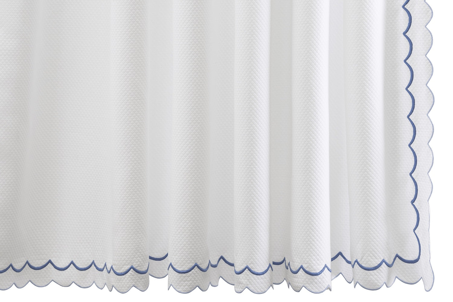 The India Pique Shower Curtain Collection by Matouk features a white cotton blend curtain with a subtle diamond pique textured pattern, elegantly finished with blue India double-scallop embroidery along the bottom and right edge.