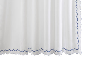 The India Pique Shower Curtain Collection by Matouk features a white cotton blend curtain with a subtle diamond pique textured pattern, elegantly finished with blue India double-scallop embroidery along the bottom and right edge.