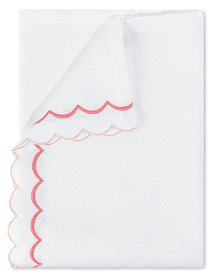 A neatly folded napkin from the Matouk India Pique Bedding Collection in Blush, featuring a white textured cotton blend with diamond pique and pink and red double-scallop embroidery.